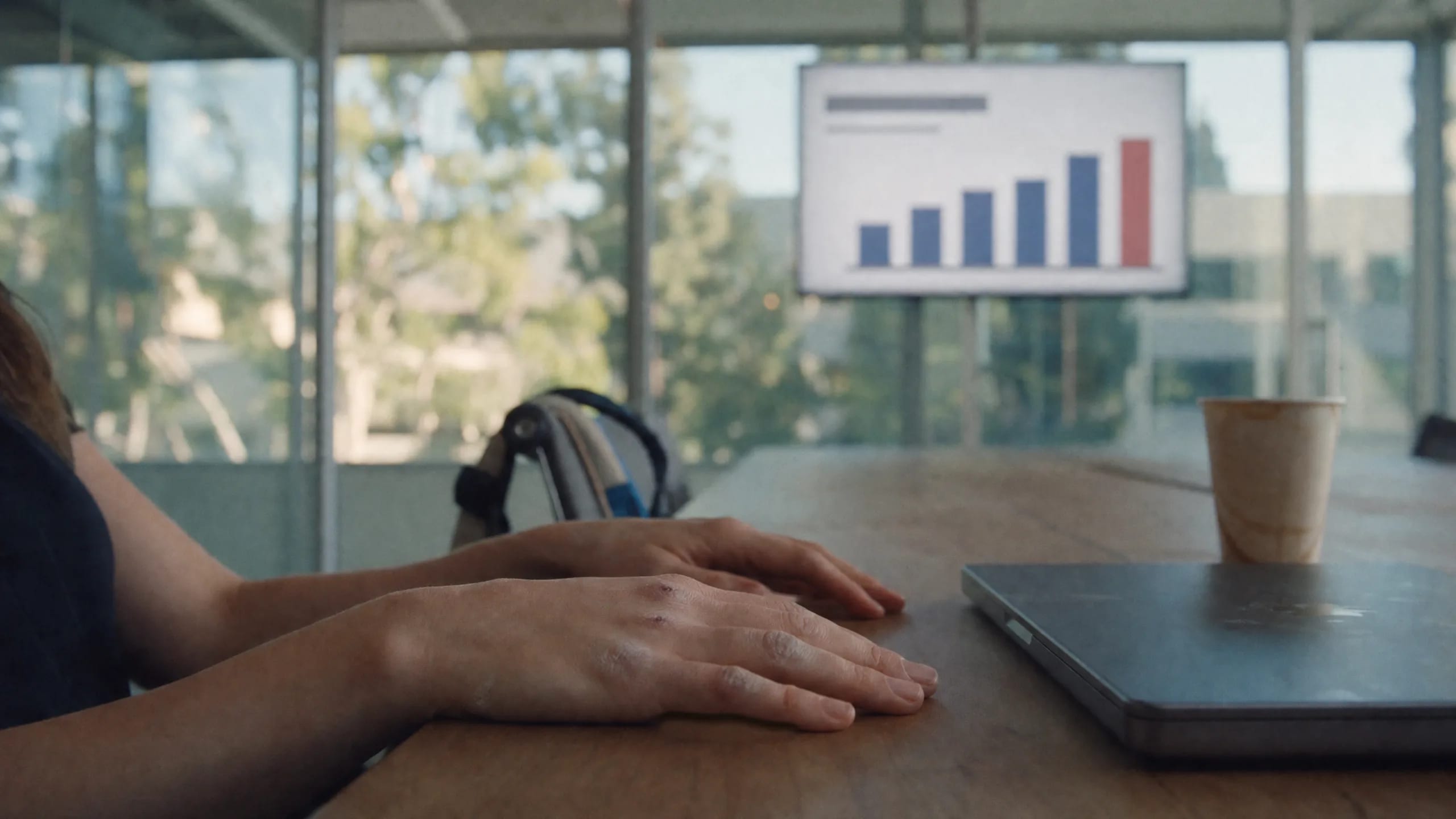 A woman's relaxed hands on a meeting-room table, her slides with a bar chart up on the wall screen behind, a gym bag strap on the chair