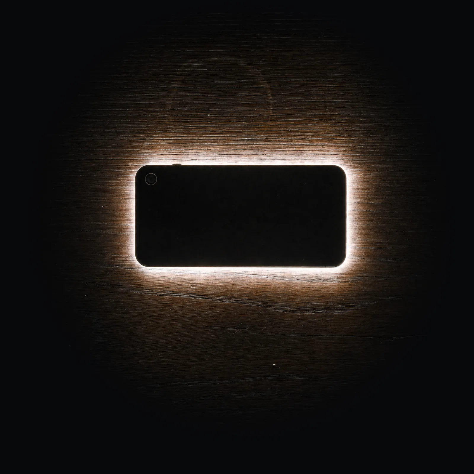 Seen from above in a dark room, a phone lies face down on a walnut table. Its screen is on, and the only light is what escapes around its edges.